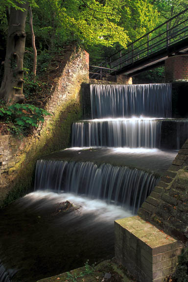 WAL: Northern Region, Flintshire County, Clwydian Hills, Holywell, Greenfields Valley Heritage Park, Water cascades over a mill pond's brick weir, in these ruins from an 18th C. copper rolling plant, now in a forest. [Ask for #246.071.]