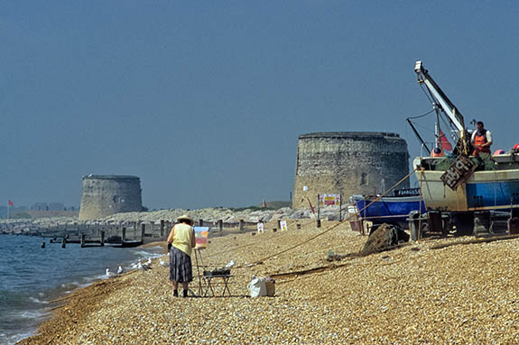 ENG: South East Region, Kent, Romney Marsh, Romney Marsh Beaches, Fishing boats drawn up on a shingle beach; woman artist is painting them; Martello Towers in the bkgd; Hythe Artillery Range visible [Ask for #256.505.]