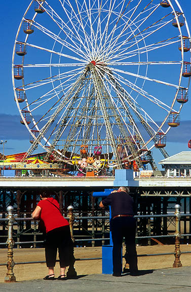 ENG: Blackpool Borough, Southern Beachfront, The Golden Mile, View past Victorian iron rails along top of seawall, across low tide sands, to Ferris wheel on pier; a couple looks through pay telescope [Ask for #262.020.]